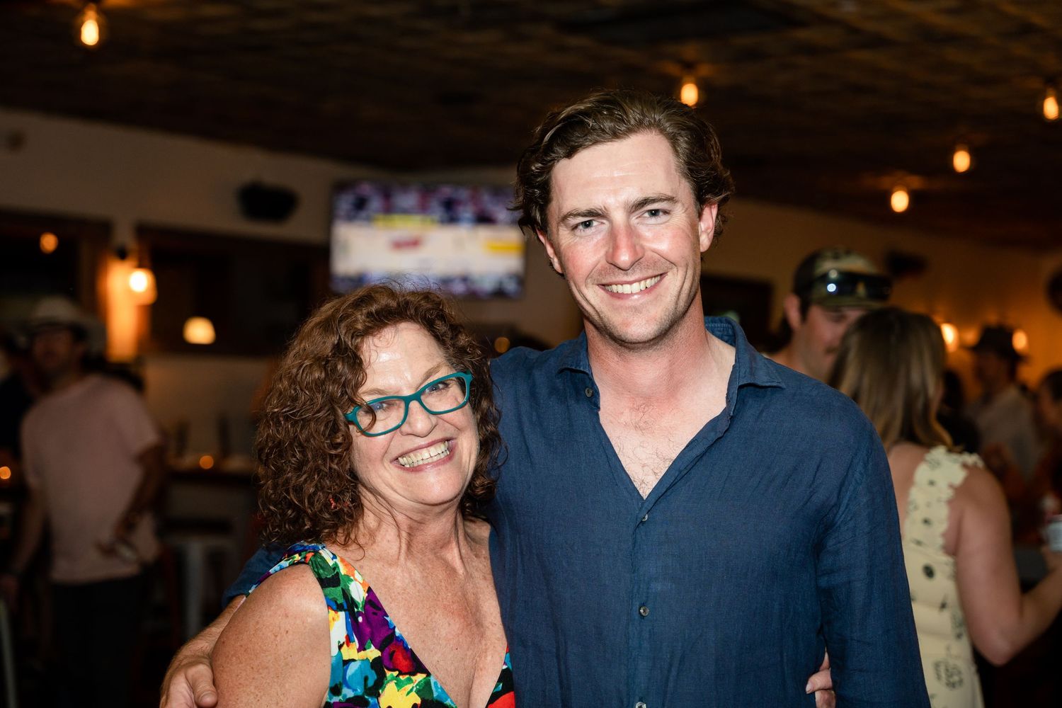Two people smiling together at an indoor social gathering with warm lighting.