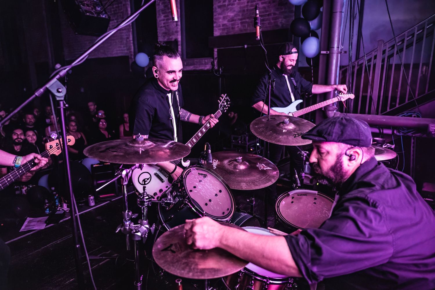 Musicians performing on stage with purple lighting and drum set visible.
