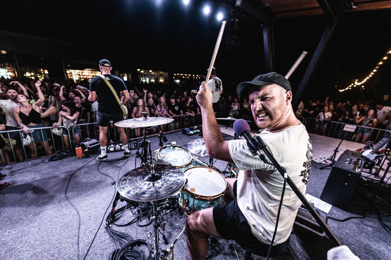 Drummer performing energetically behind drum kit on brightly lit concert stage.