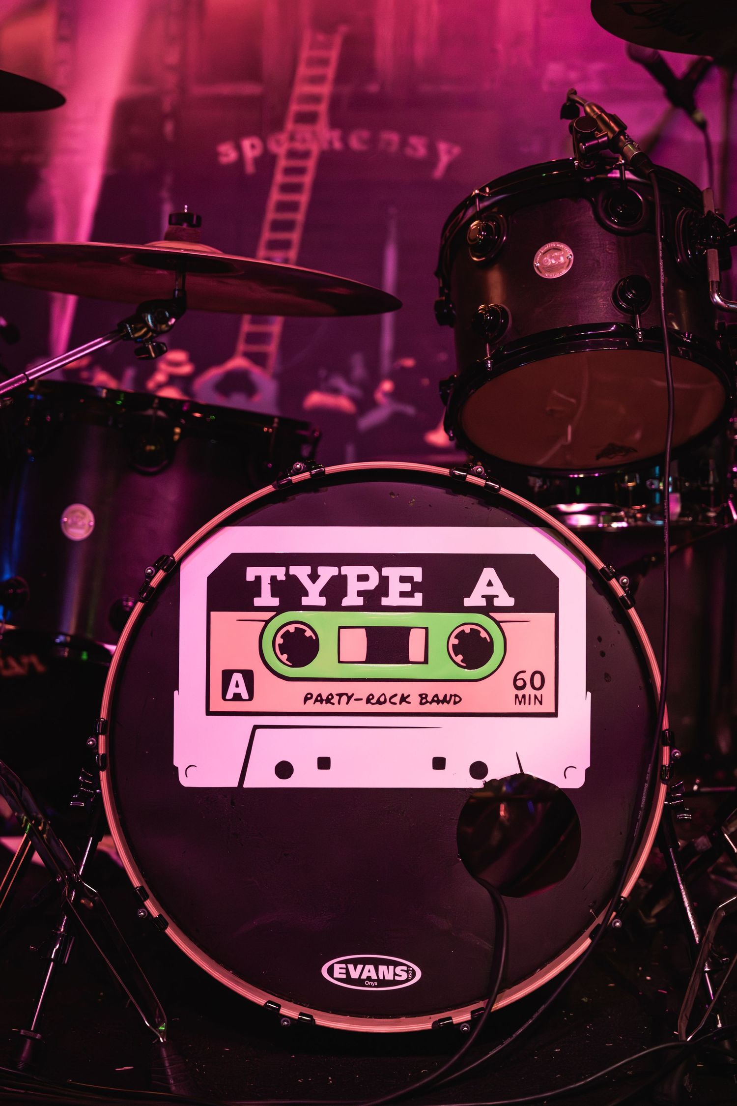A drum kit with a custom bass drum head featuring a Type A cassette tape logo illuminated in pink lighting.