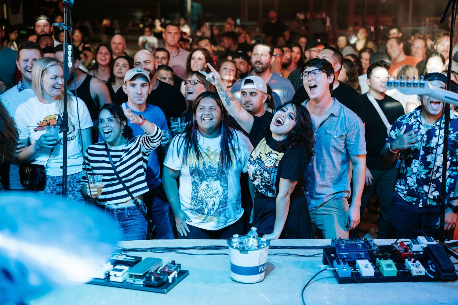 Concert crowd watches enthusiastically as performers play music in a dimly lit venue with blue lighting.