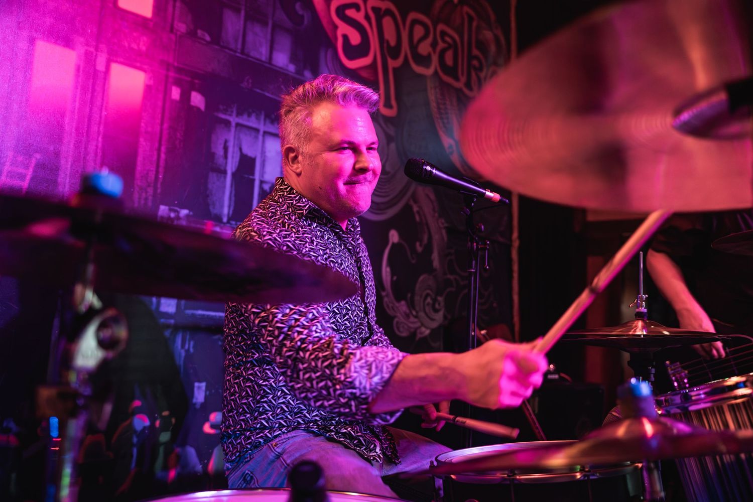 Dynamic shot of drummer performing with colorful magenta stage lighting.