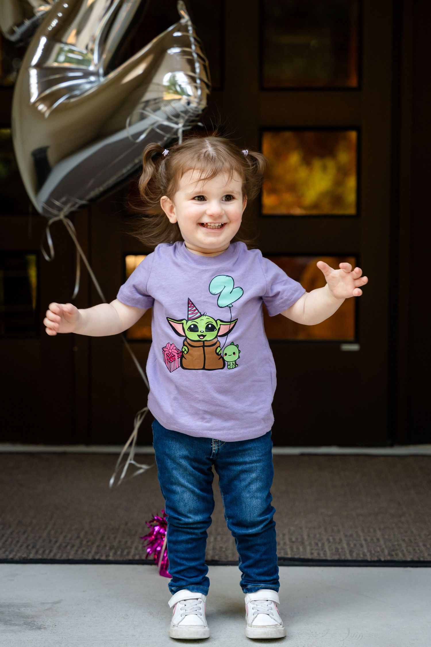 An excited child in a Baby Yoda shirt poses with silver balloons against a dark doorway.
