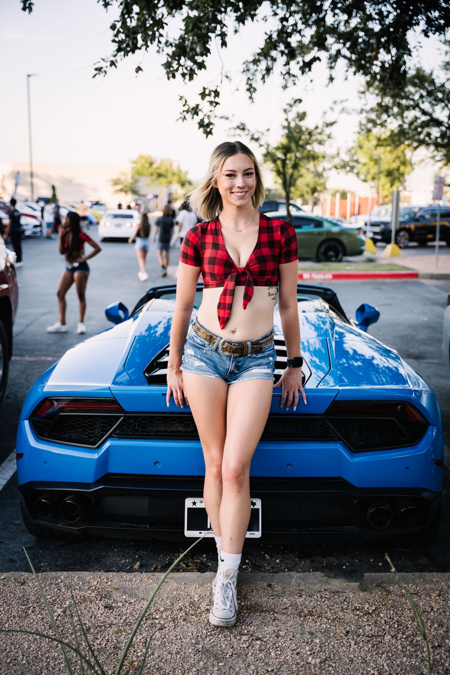 Model in red plaid top sits on blue Lamborghini hood wearing denim shorts.