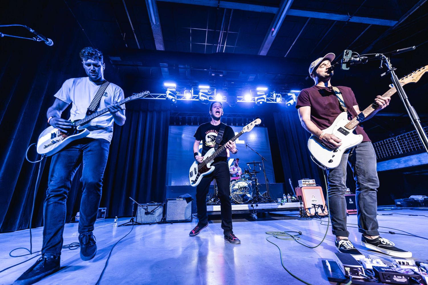 Four musicians perform on stage with guitars under blue stage lighting at a live concert venue.