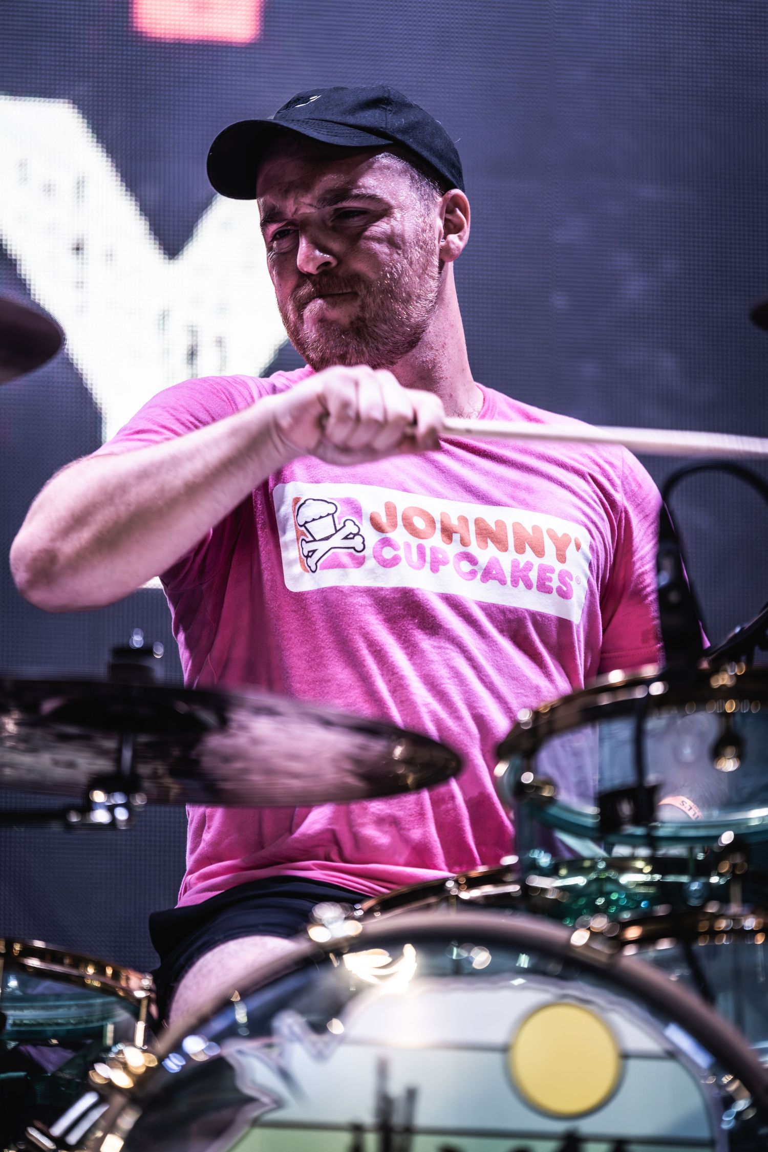 Drummer in pink Johnny Cupcakes t-shirt performing energetically on stage during live concert performance.