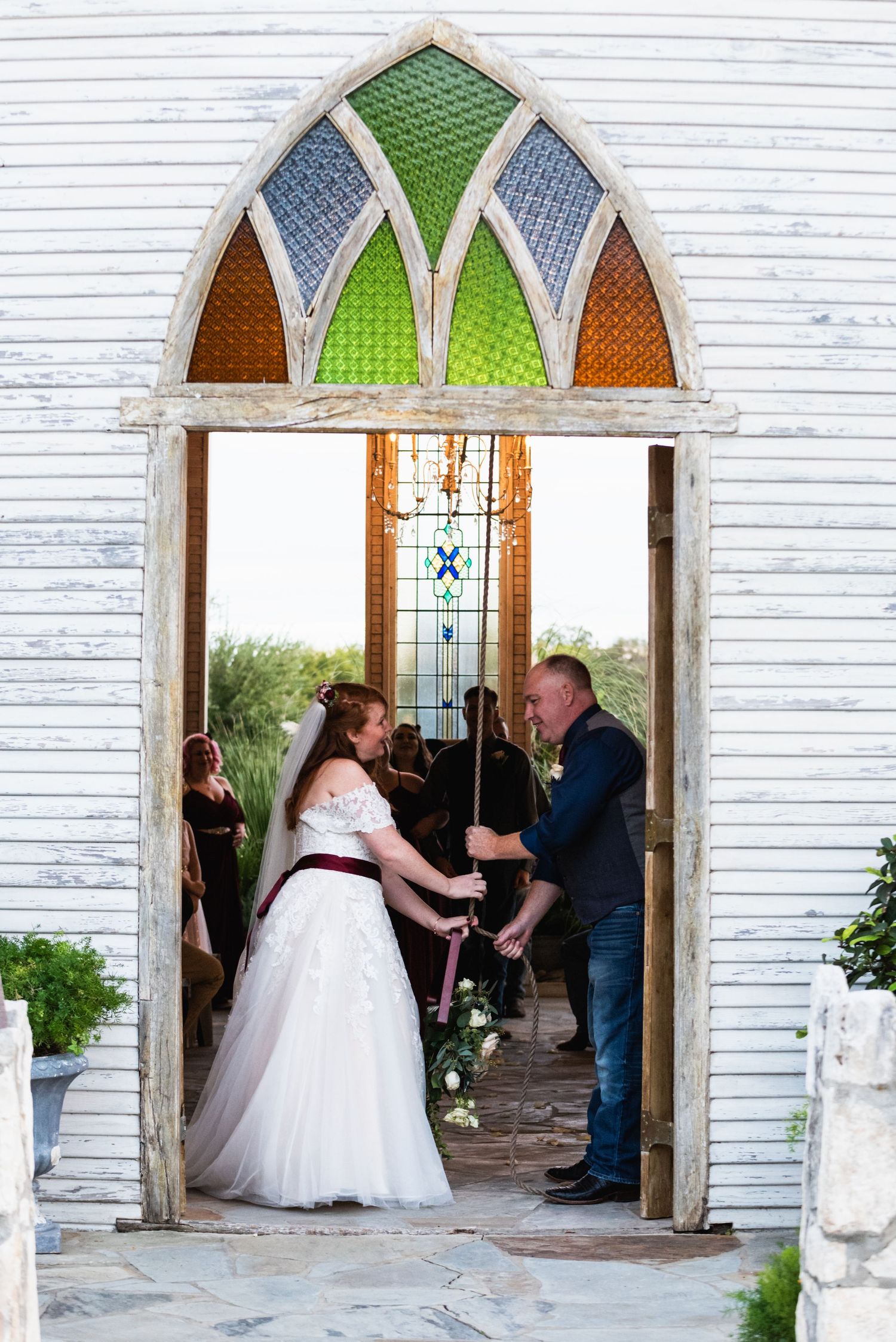 A stained glass arch frames a couple during their wedding ceremony in a rustic white chapel.