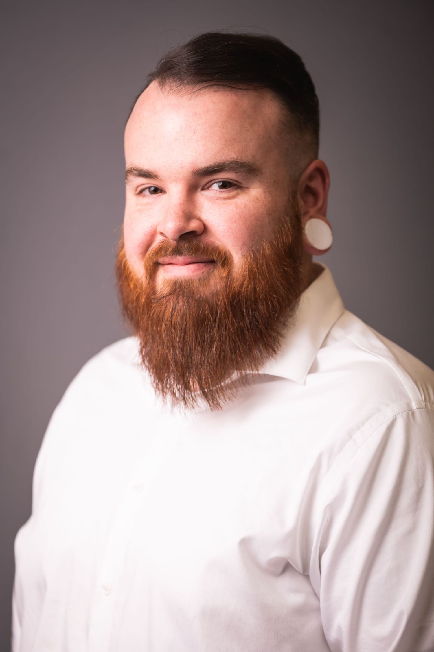 Portrait of person with red beard wearing white shirt against grey backdrop.