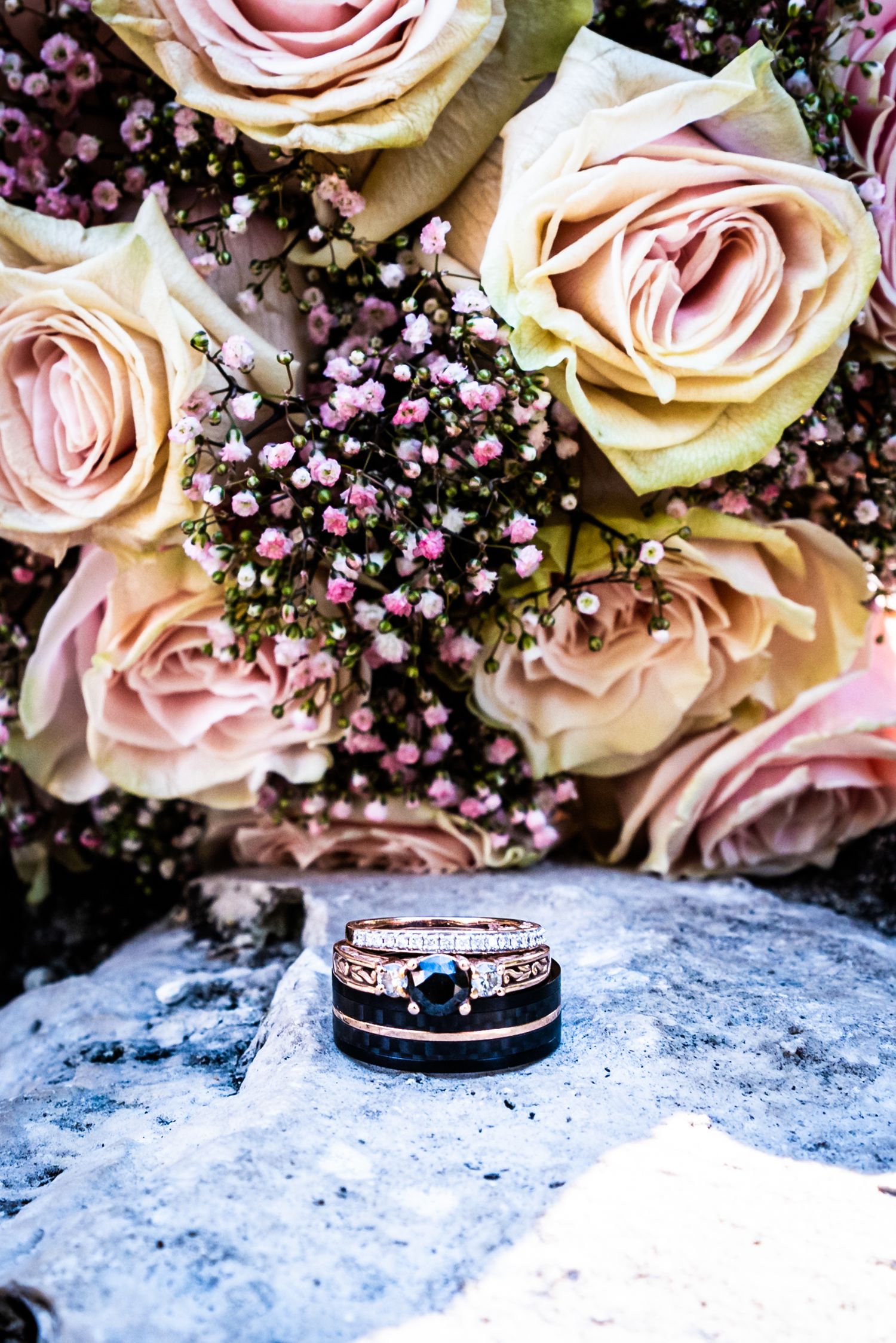Wedding rings displayed with pink roses and baby's breath flowers on a stone surface.