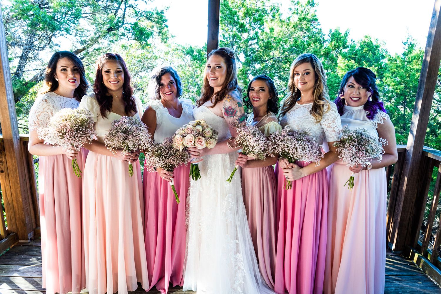 Bridesmaids in various shades of pink dresses stand together holding bouquets on an outdoor wooden platform.