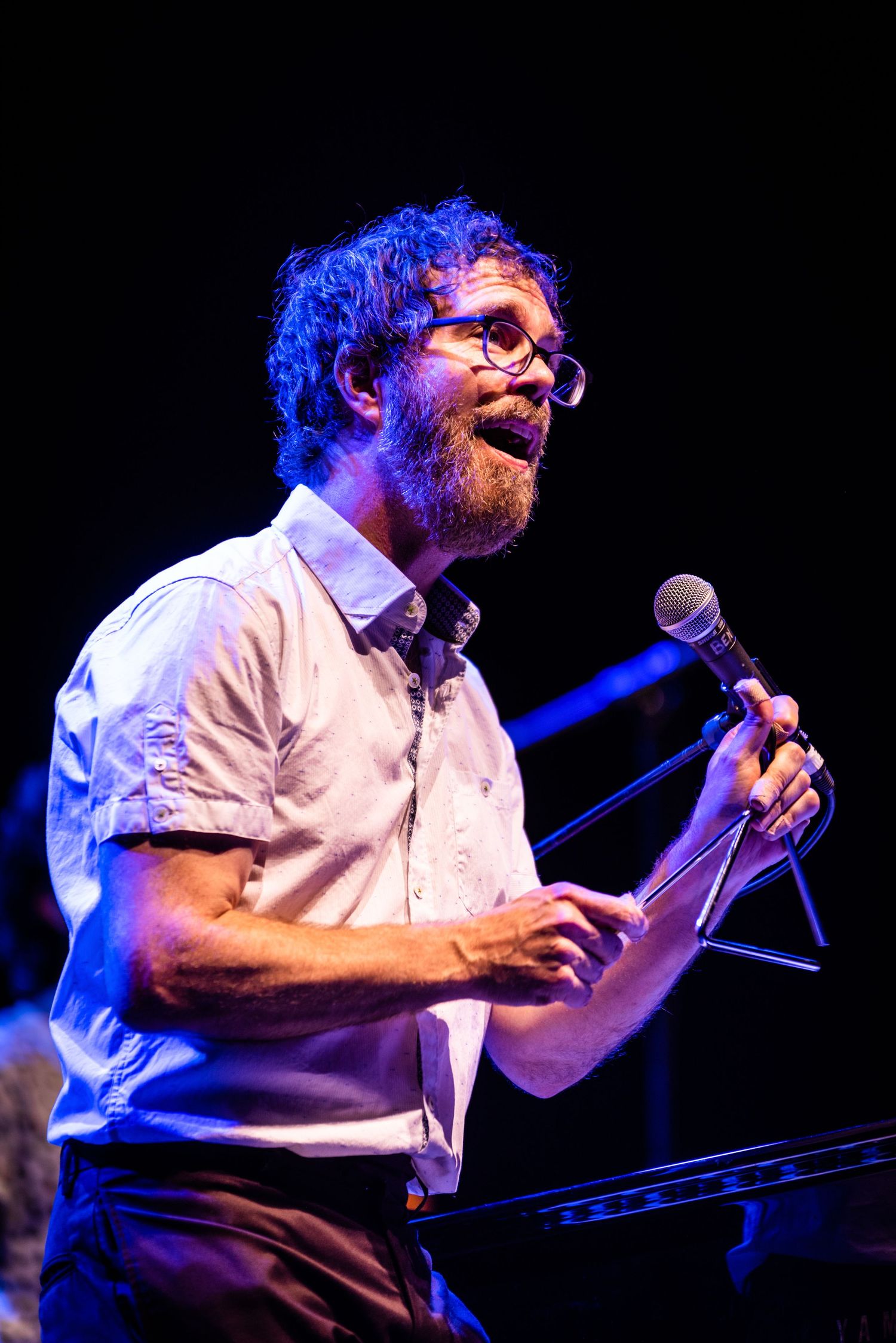 Musician performs with microphone under blue stage lighting wearing white button-down shirt.