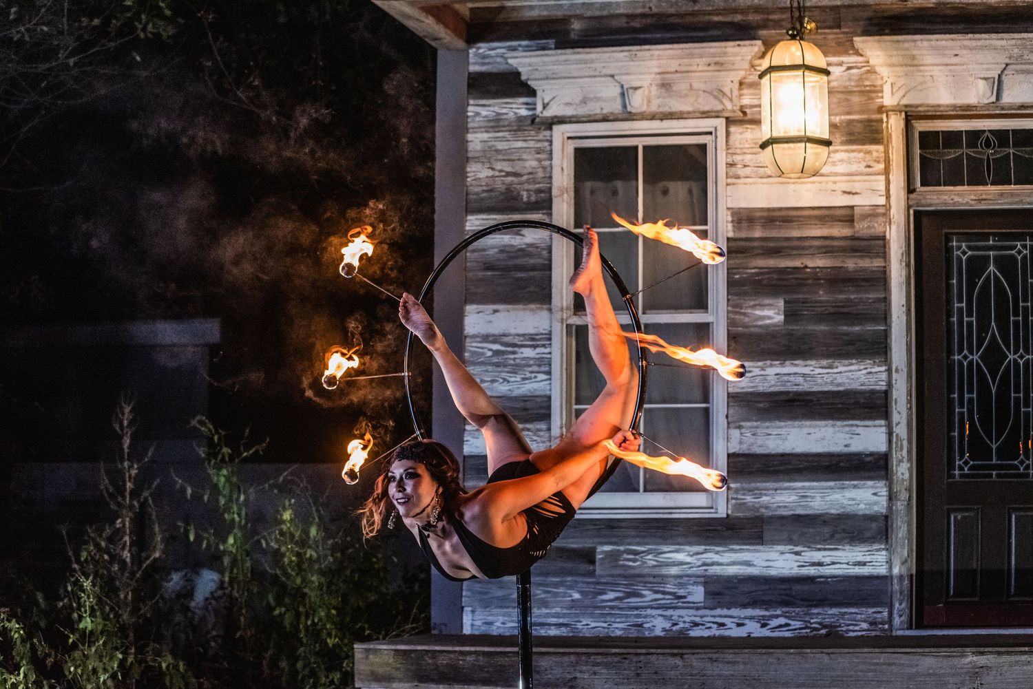 A performer in an orange outfit hangs upside down from an aerial hoop against a rustic wooden building at night.