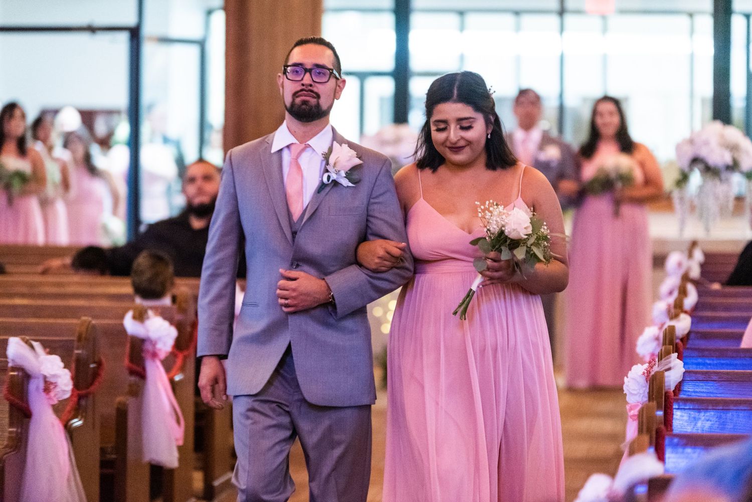 Wedding party member in grey suit escorts bridesmaid in pink down church aisle.