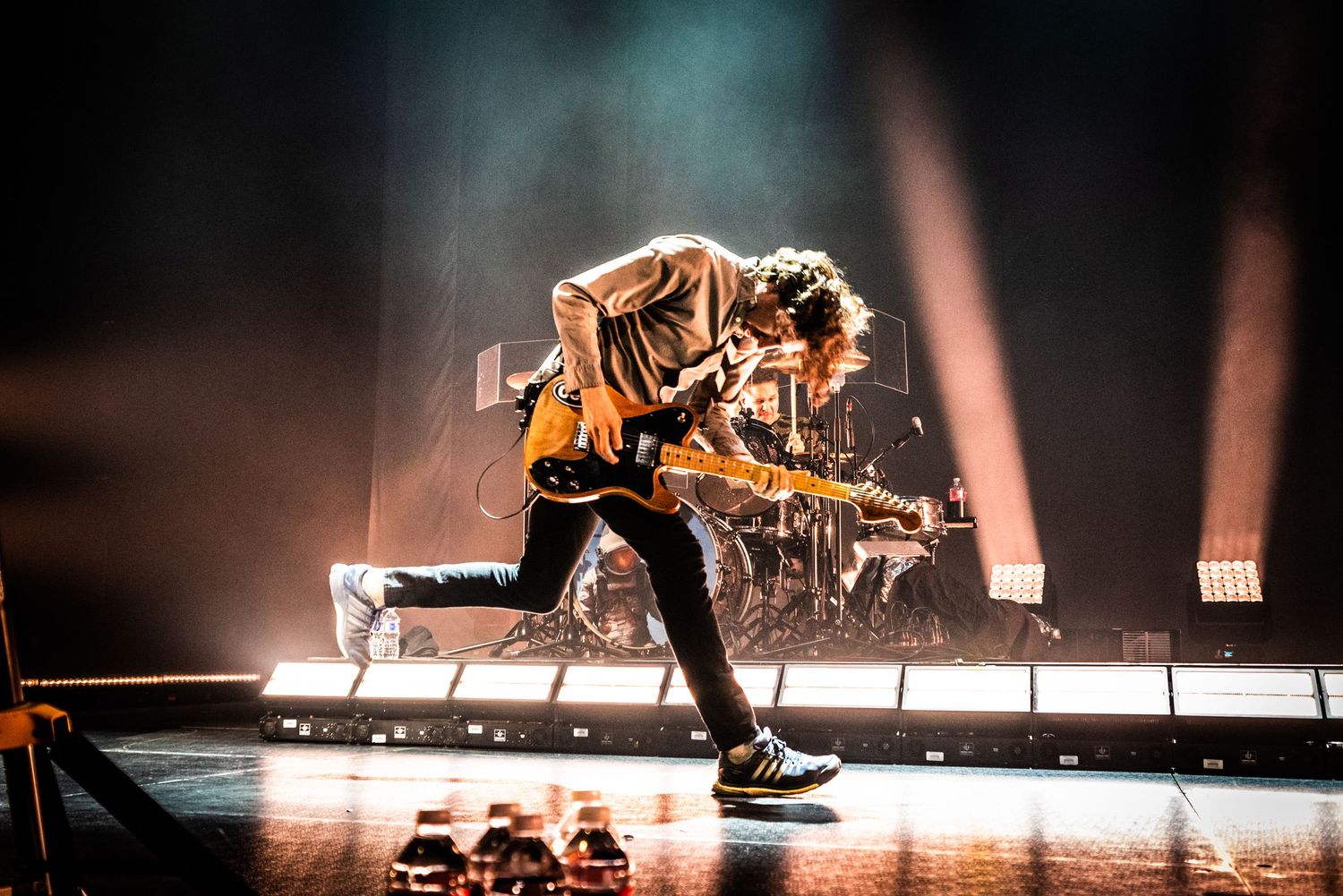 Dynamic stage performance with musician in motion against atmospheric lighting and drum kit backdrop.