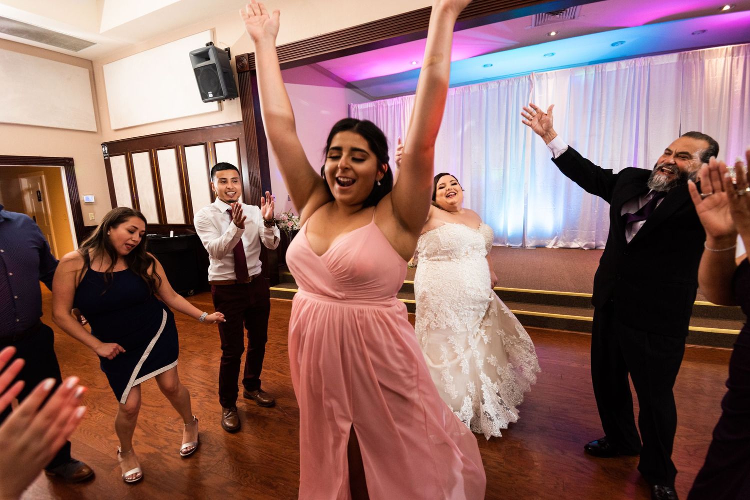 Wedding celebration with guests dancing enthusiastically under pink and purple lighting.