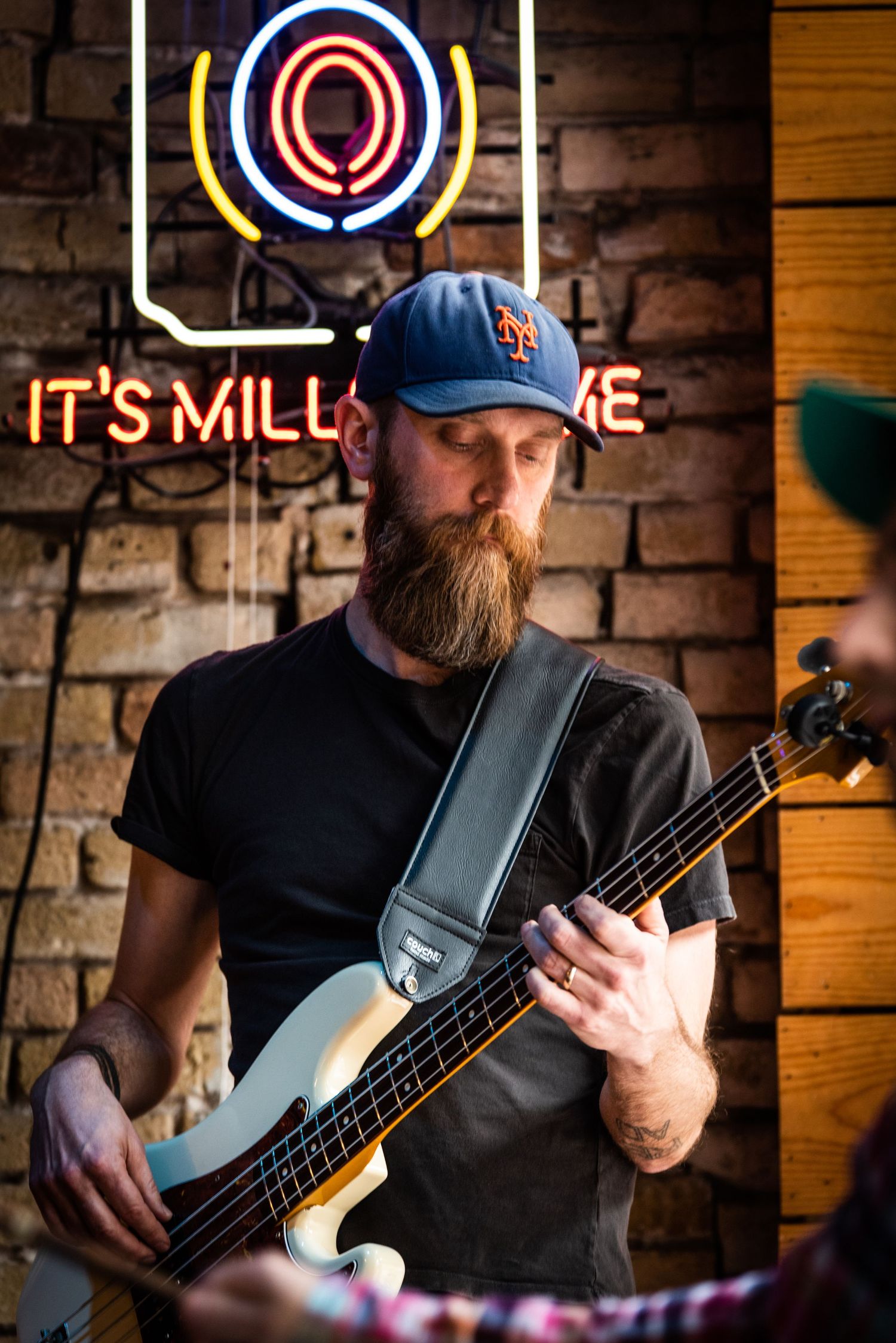 Bass player with beard performing under neon sign reading 'It's Mill' against brick wall background.