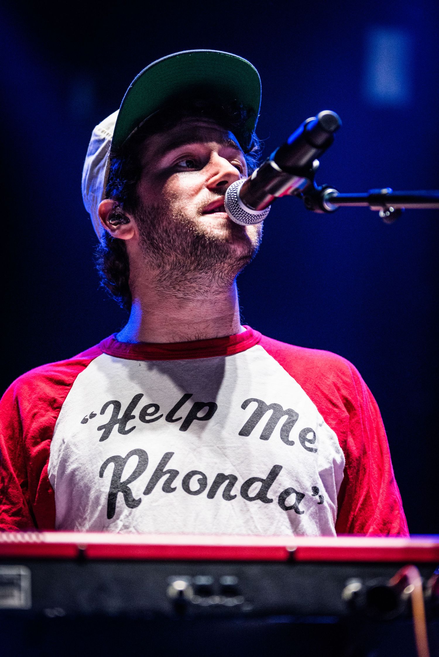 Musician in baseball cap and 'Help Me Rhonda' shirt performs at keyboard with blue stage lighting.