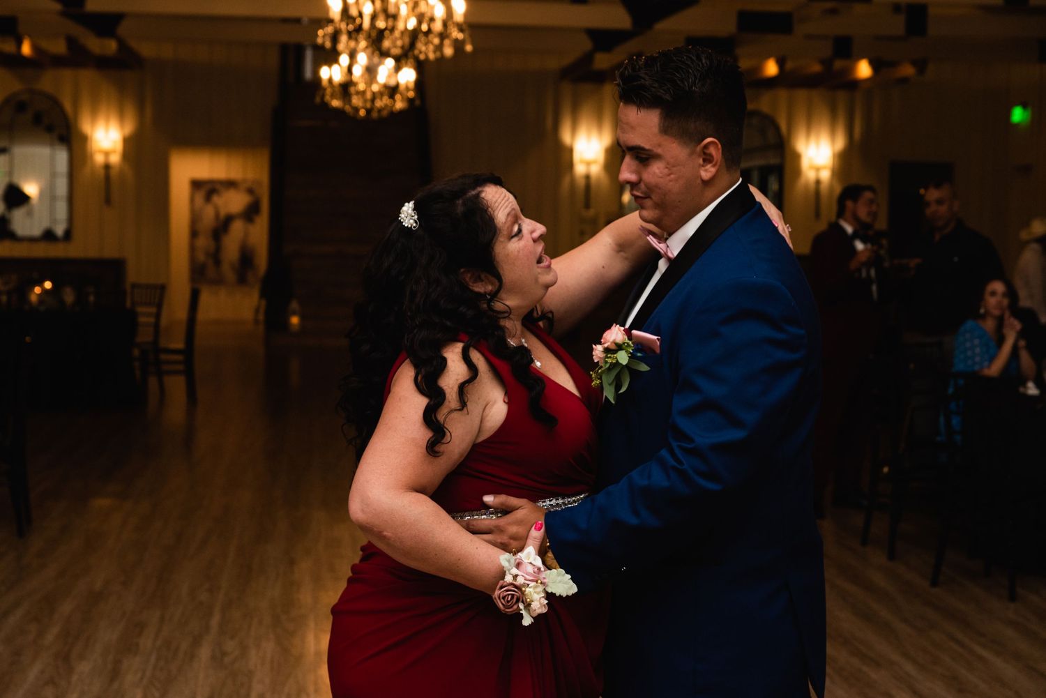 Two people dancing together at an elegant reception venue with chandeliers and hardwood floors.