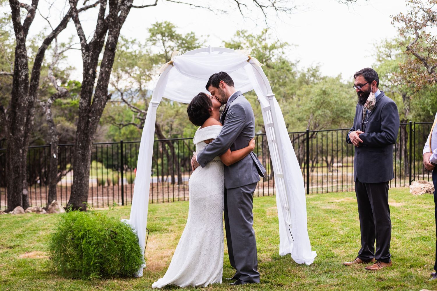 Couple embraces under white wedding arch while officiant stands nearby during outdoor ceremony.