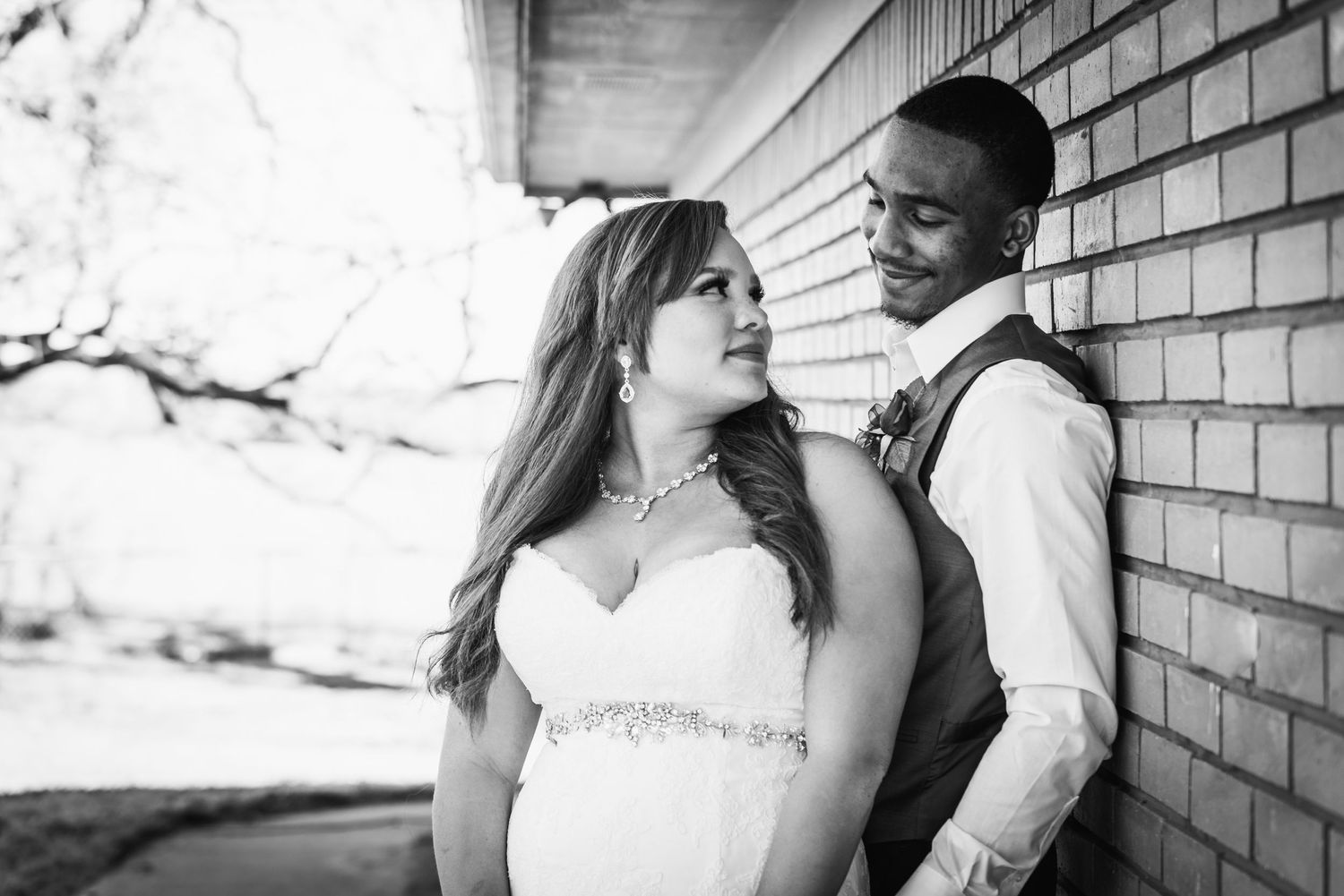Romantic black and white portrait of newlyweds posed against brick wall in elegant wedding attire.