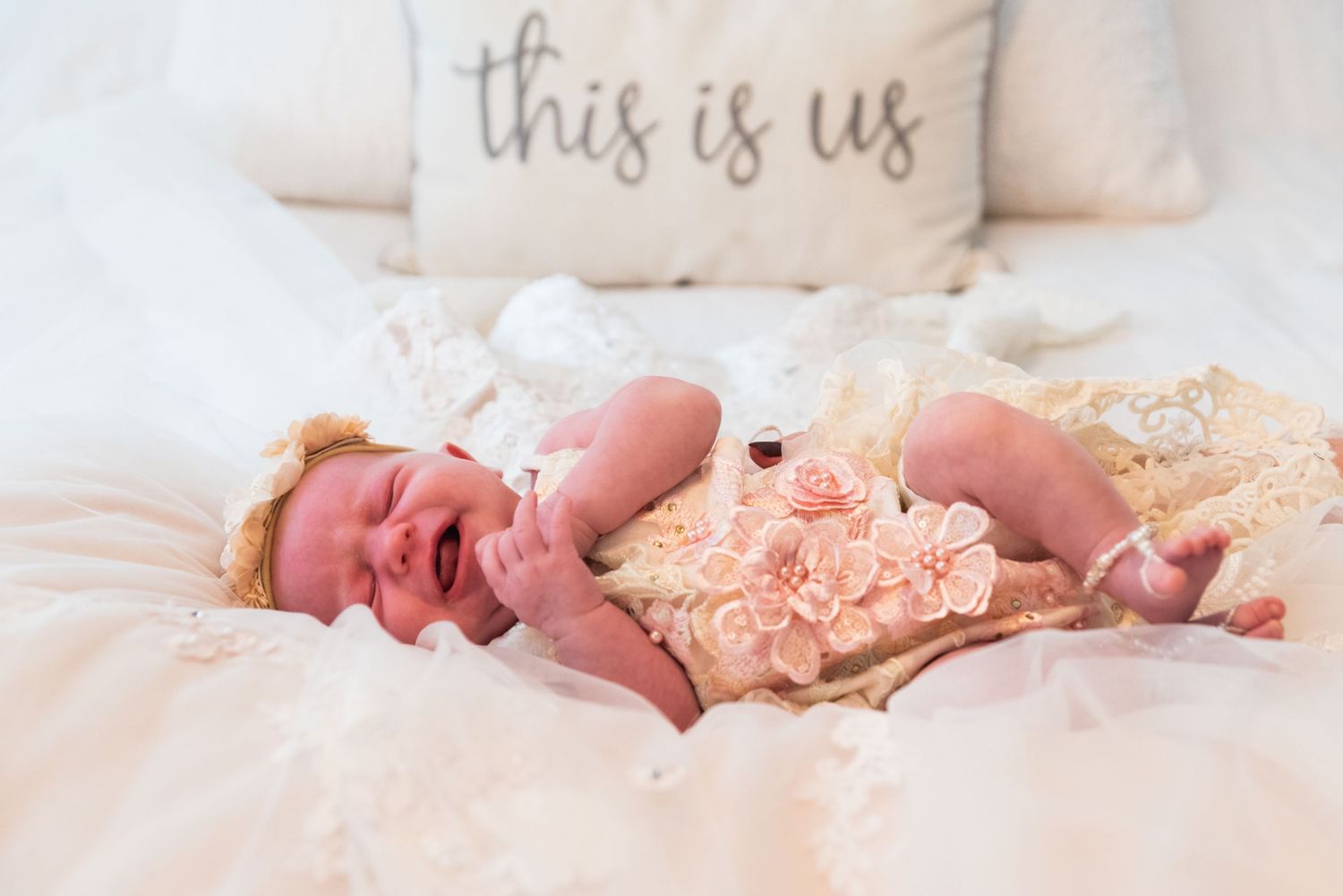 Baby lying on white bedding with decorative pillow reading 'this is us' in background.