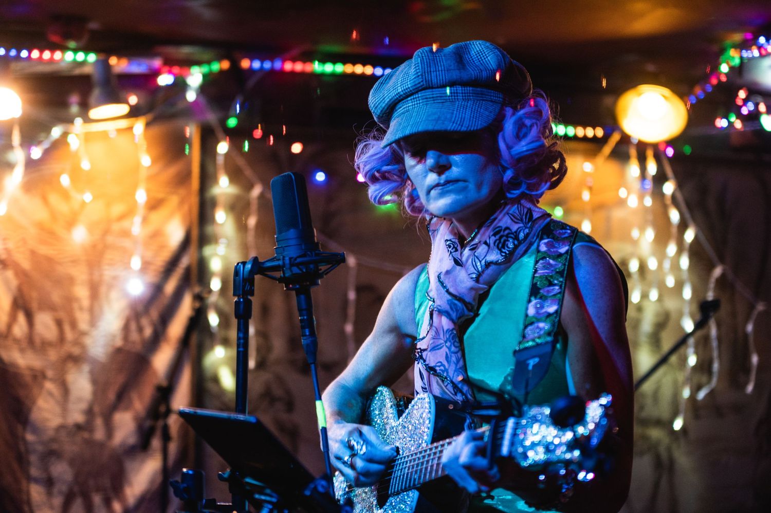 A performer with a guitar plays under blue stage lighting with string lights in the background.