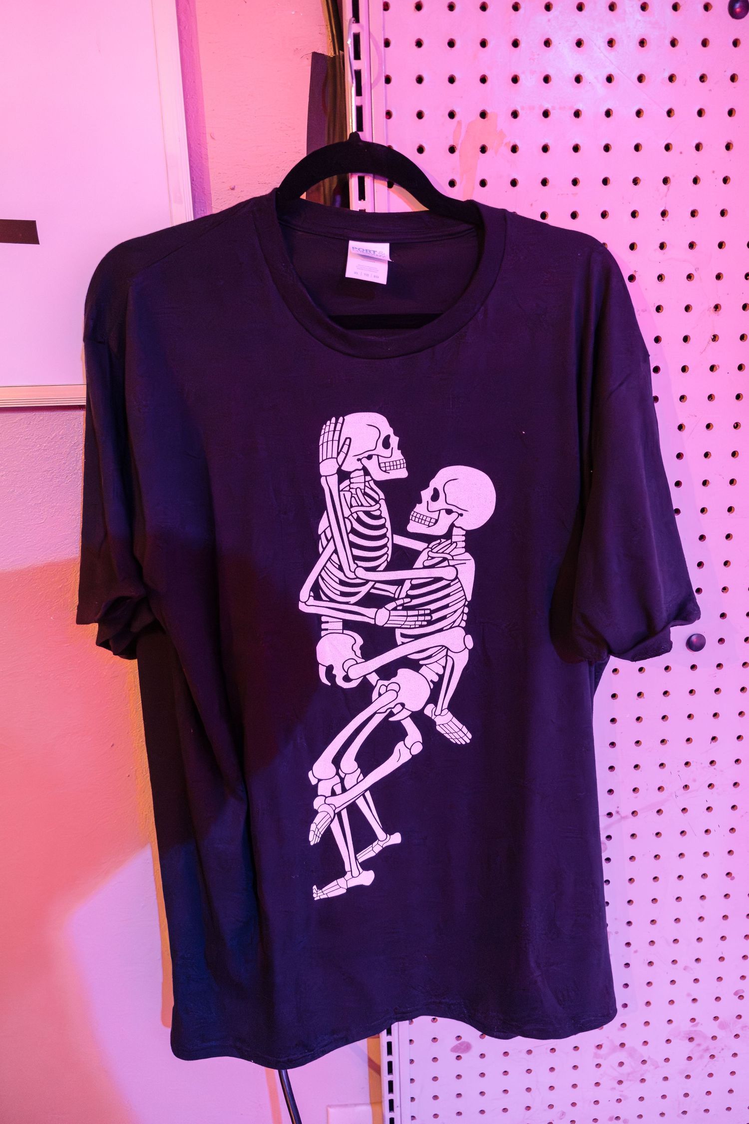 Black t-shirt with white skeleton couple design displayed on pink pegboard wall.