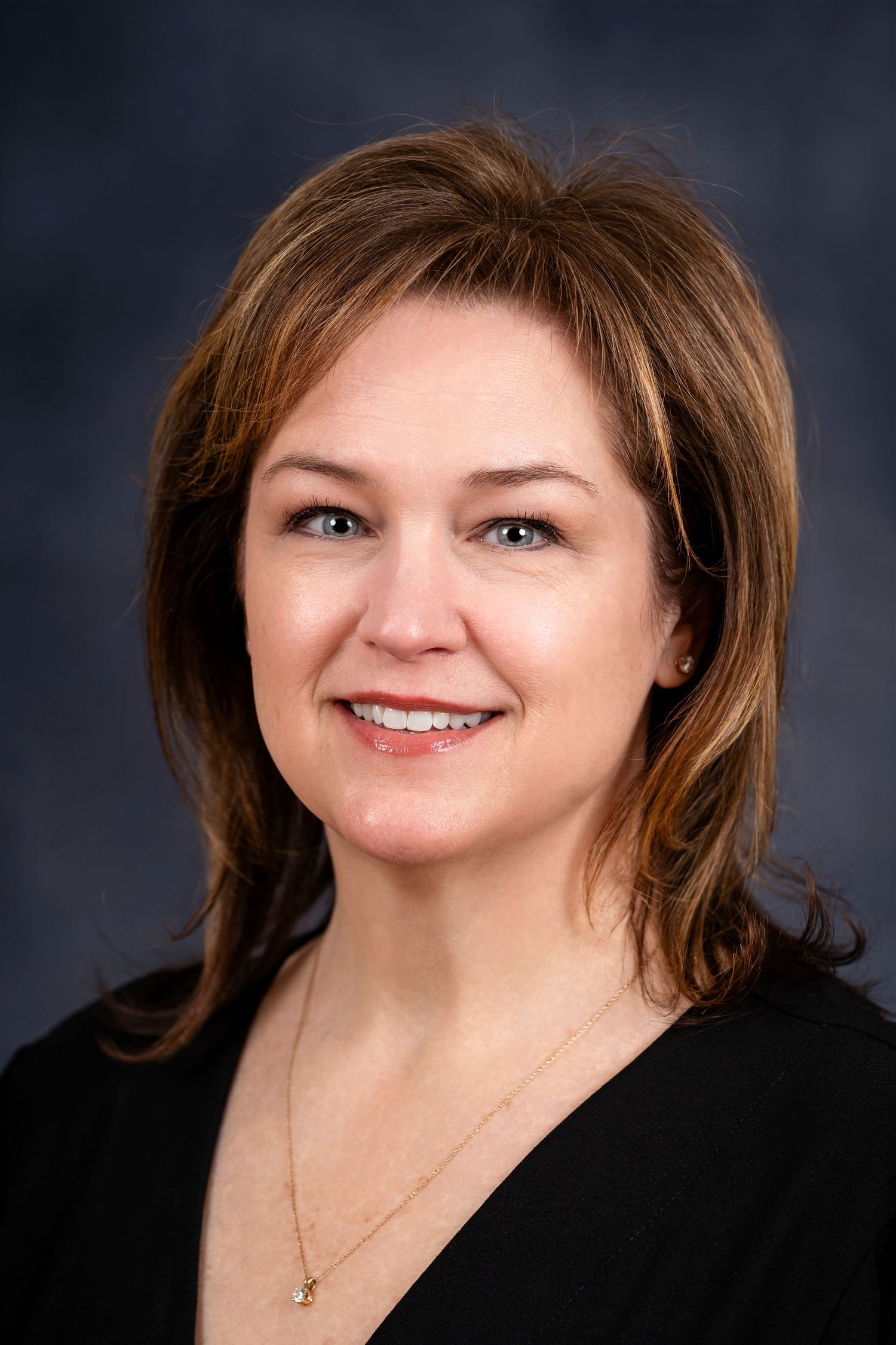 Professional headshot of a person with layered brown hair wearing a black top against dark background.