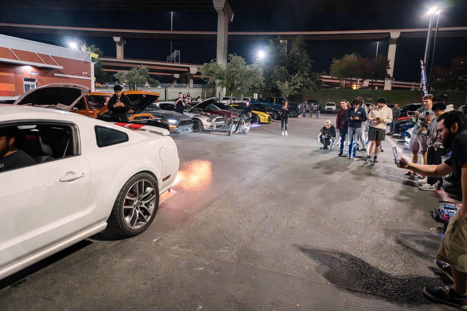 White sports car creates flame burst from exhaust during nighttime car meet.