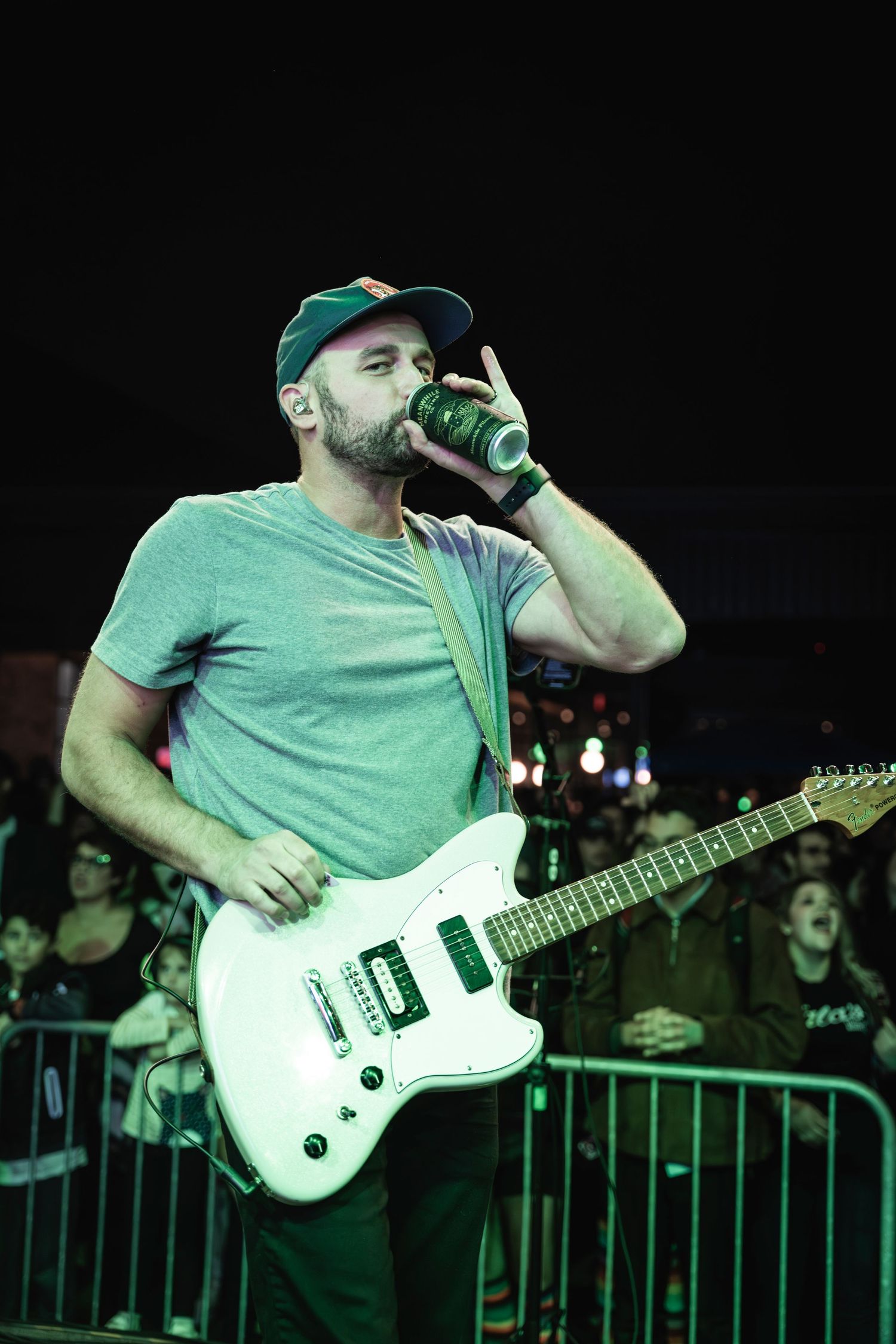 Guitarist in green shirt and cap performs while drinking from can on stage.