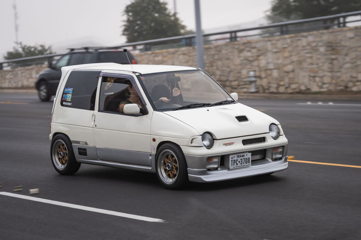 Modified white Suzuki Alto with aftermarket wheels driving on road.