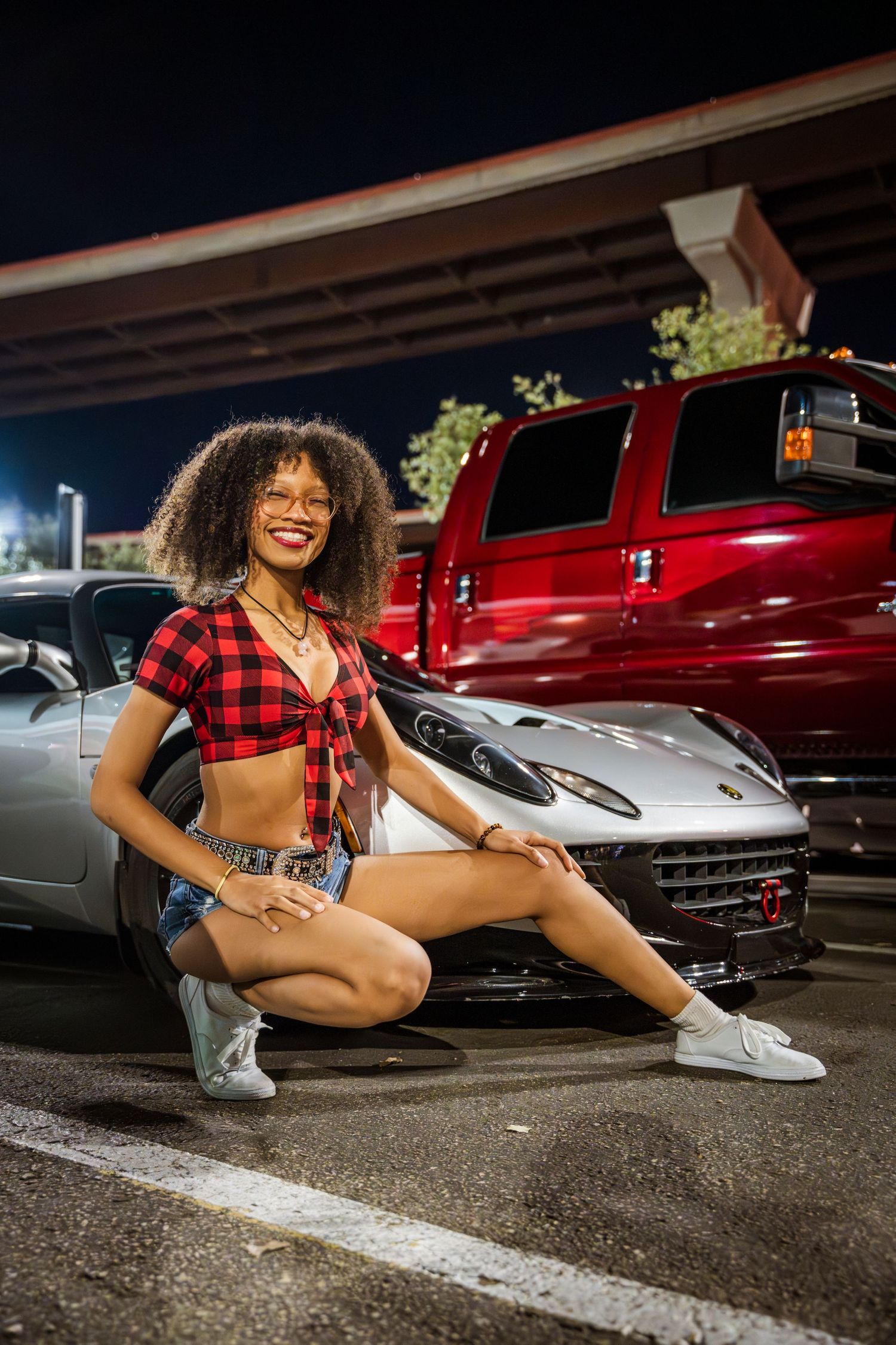 Woman in buffalo plaid crop top poses next to car in parking lot at night.