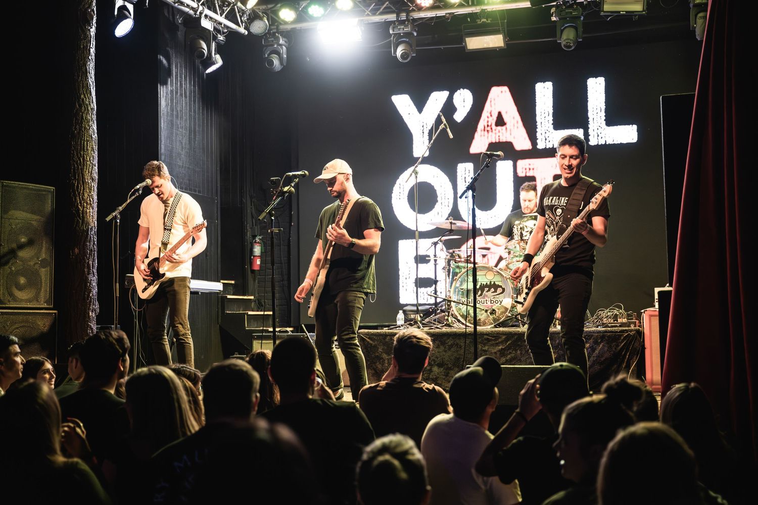 Band performs on stage with 'Y'ALL OUT' sign backdrop while crowd watches at energetic rock concert.