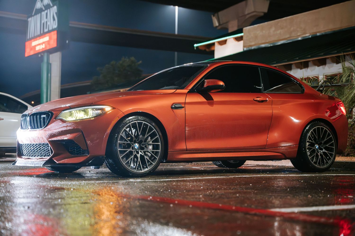 Orange BMW M2 Competition sports car parked on wet asphalt at night.
