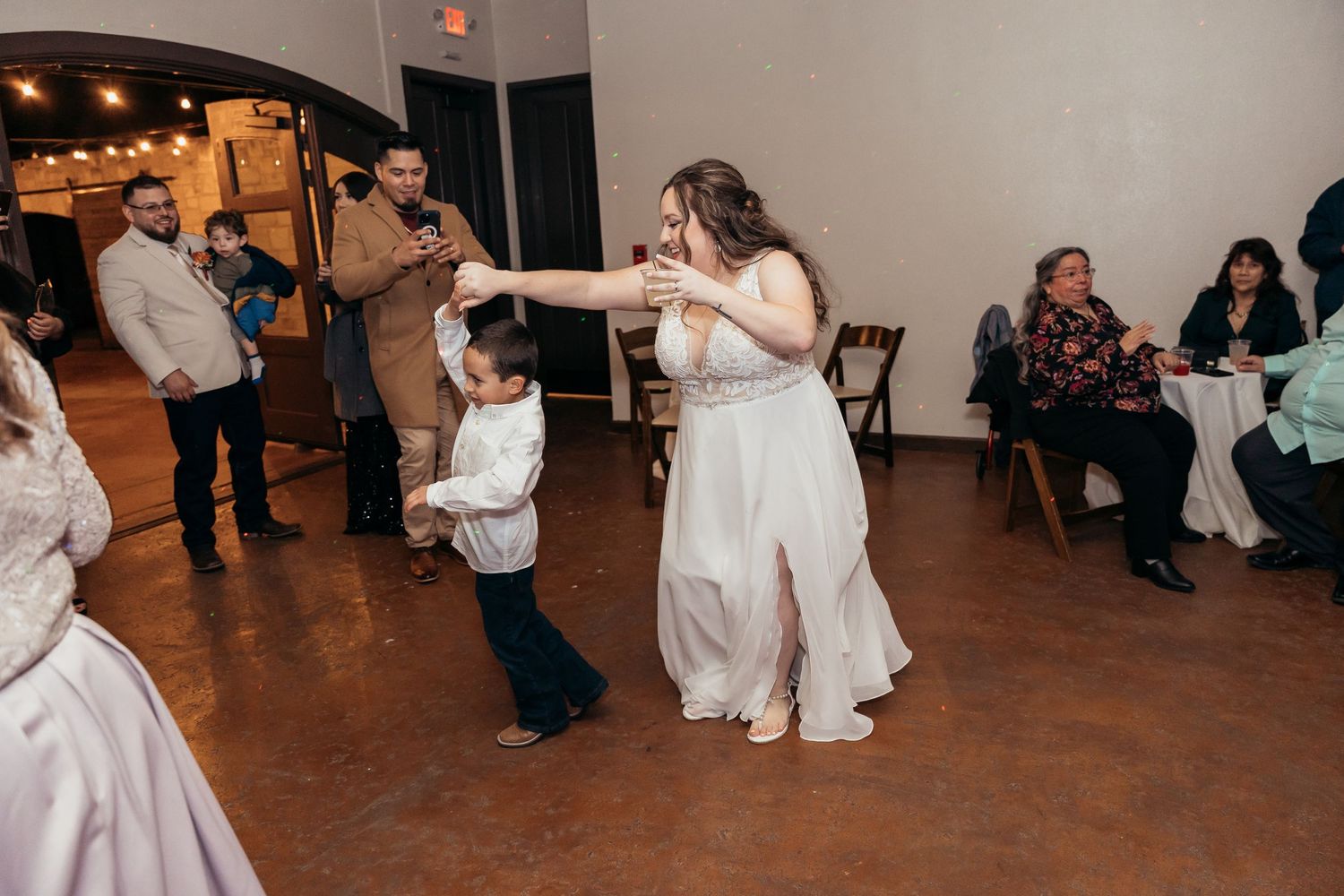 Bride in a white dress dances with a young boy during a wedding reception on a wooden dance floor.