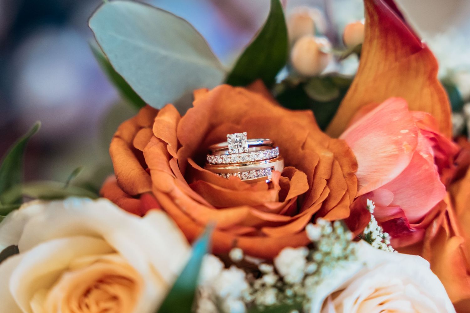 Diamond wedding rings nestled in an orange rose among mixed floral bouquet.