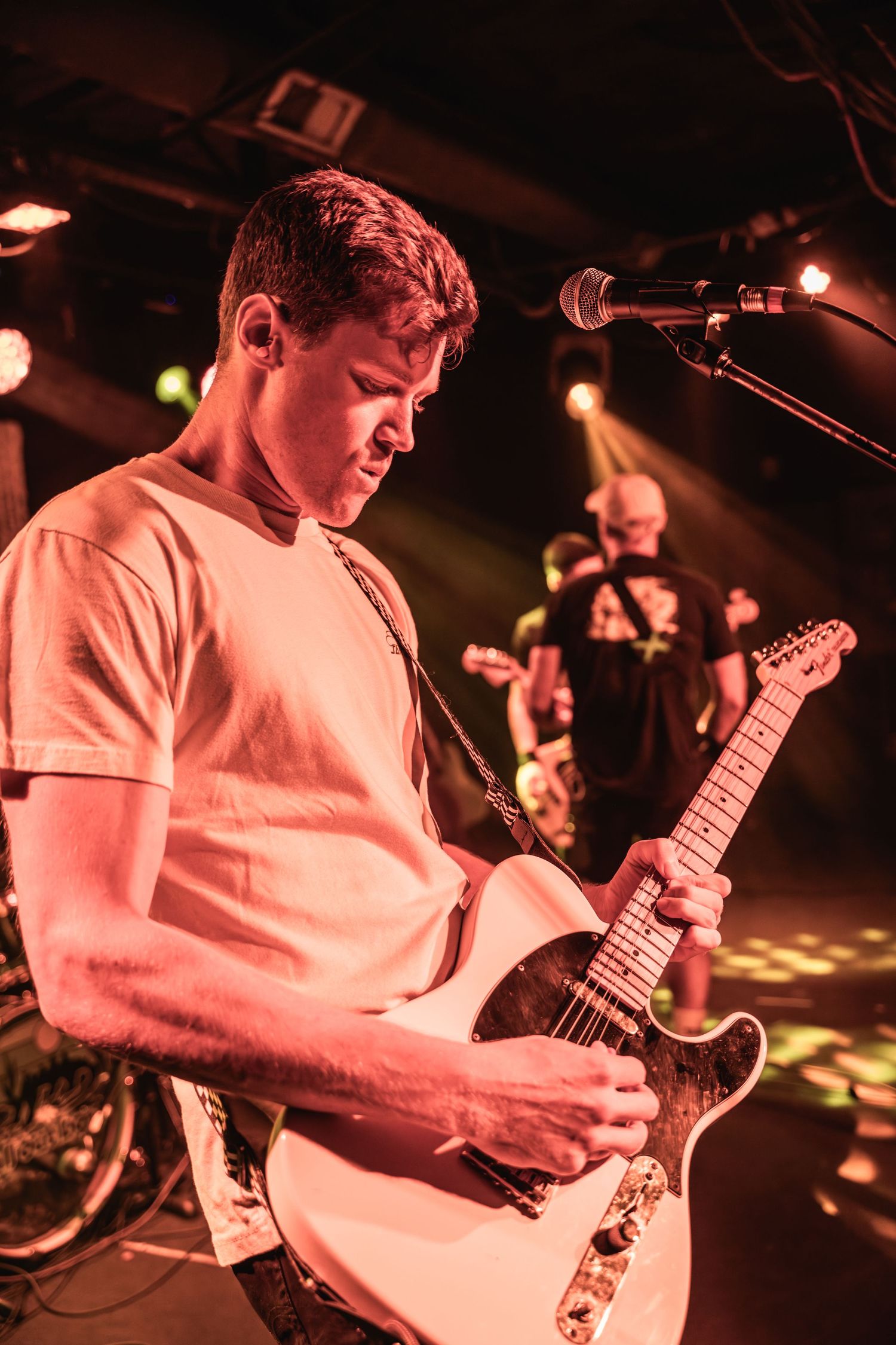 Guitarist performing onstage in a white t-shirt illuminated by warm red lighting during live concert performance.
