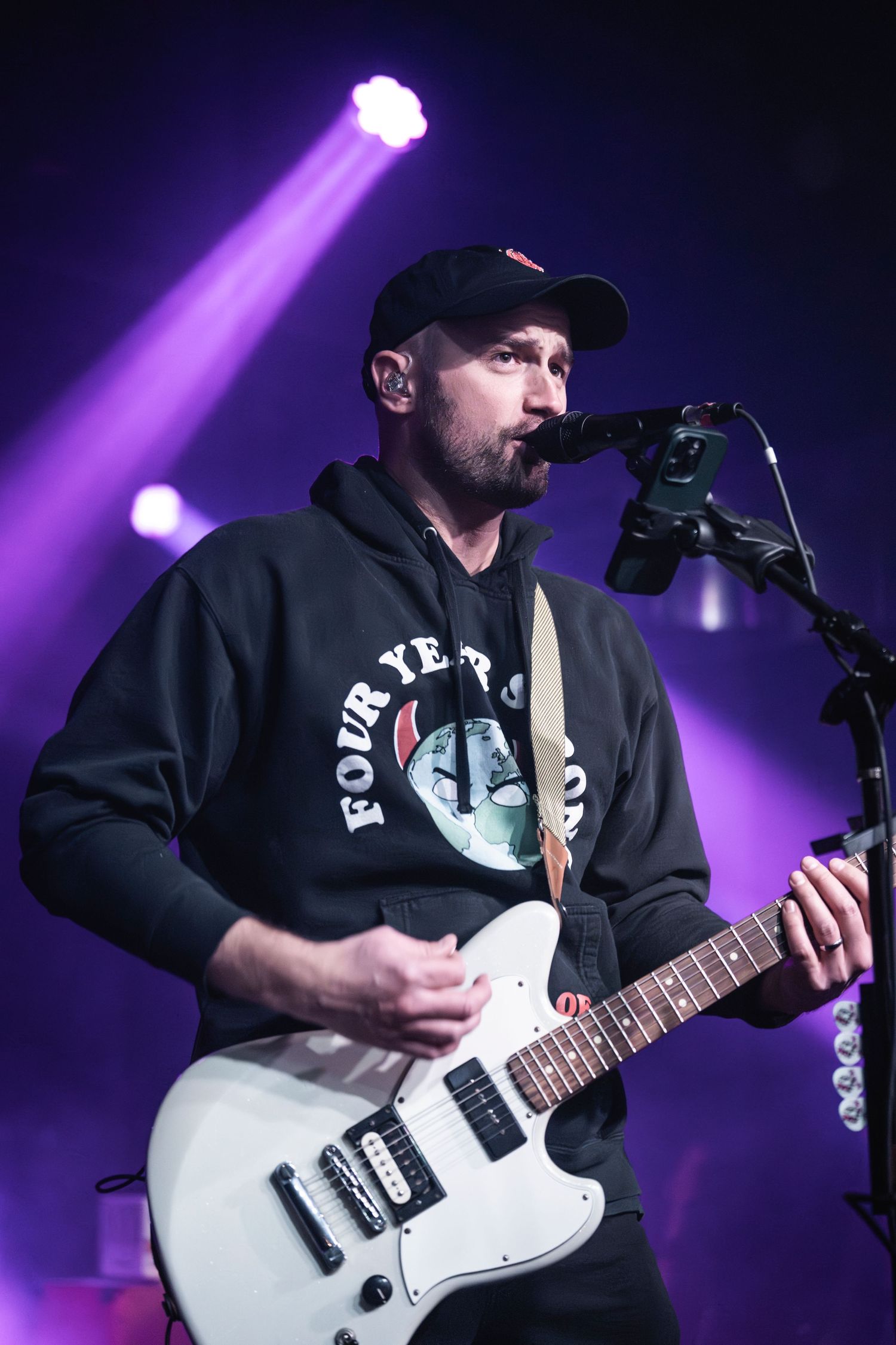 Musician in black hoodie performs with white electric guitar under purple stage lighting.