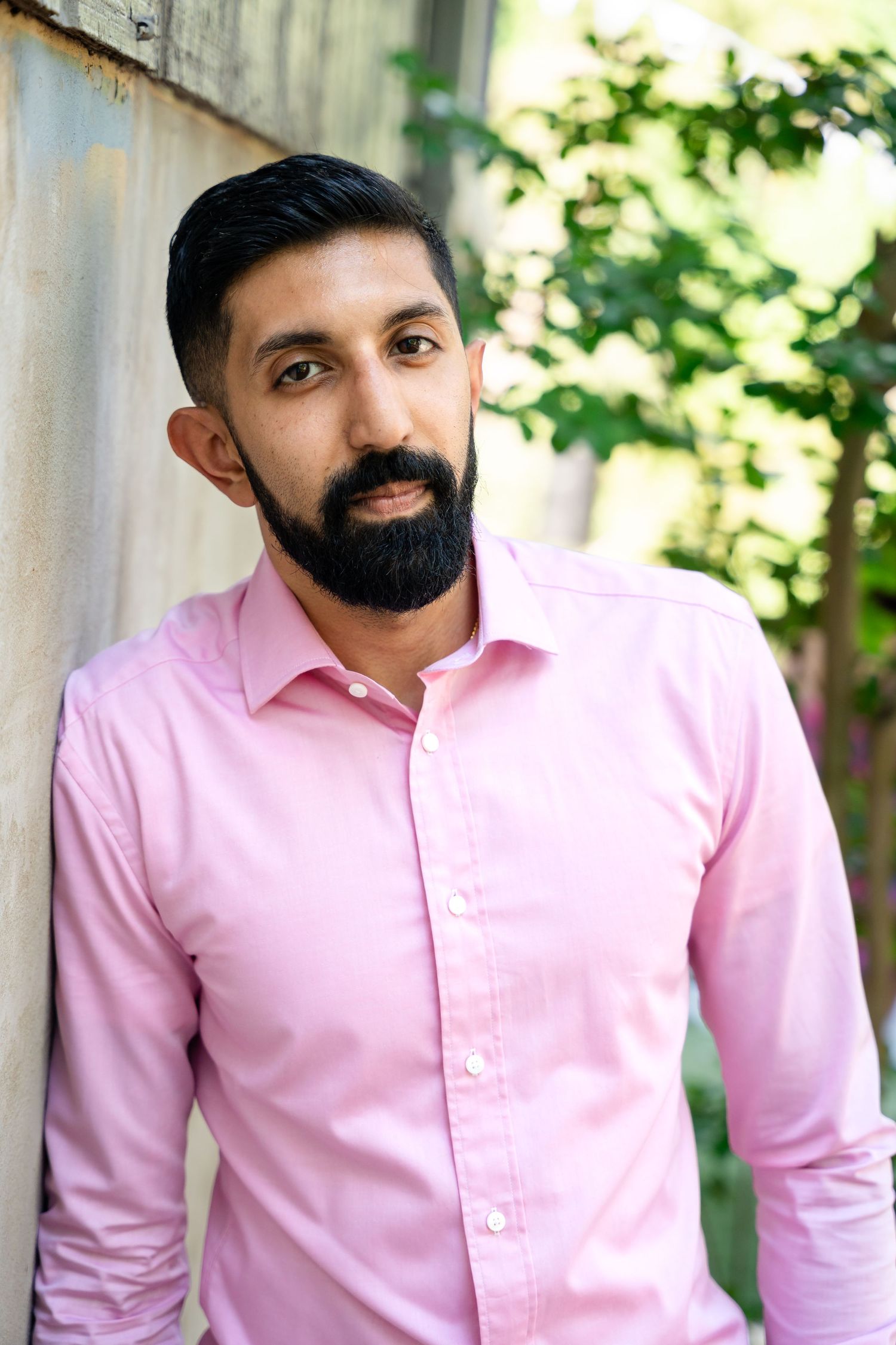 A portrait of someone wearing a pink button-down shirt against a textured wall backdrop.