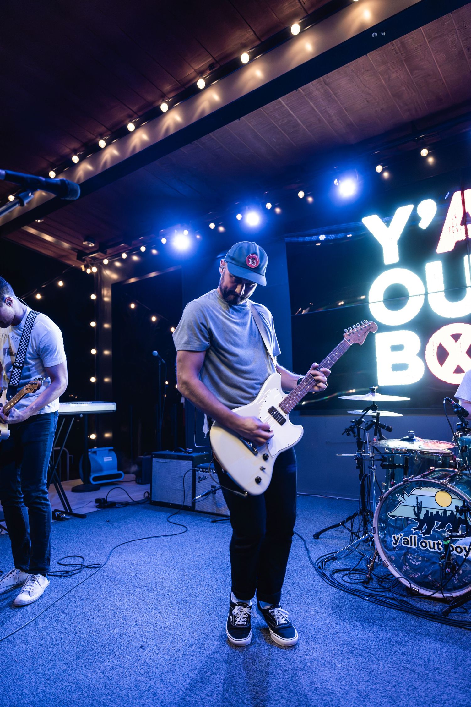 Guitarist performing on stage with blue lighting and 'Y'ALL OUT BOY' text in background.