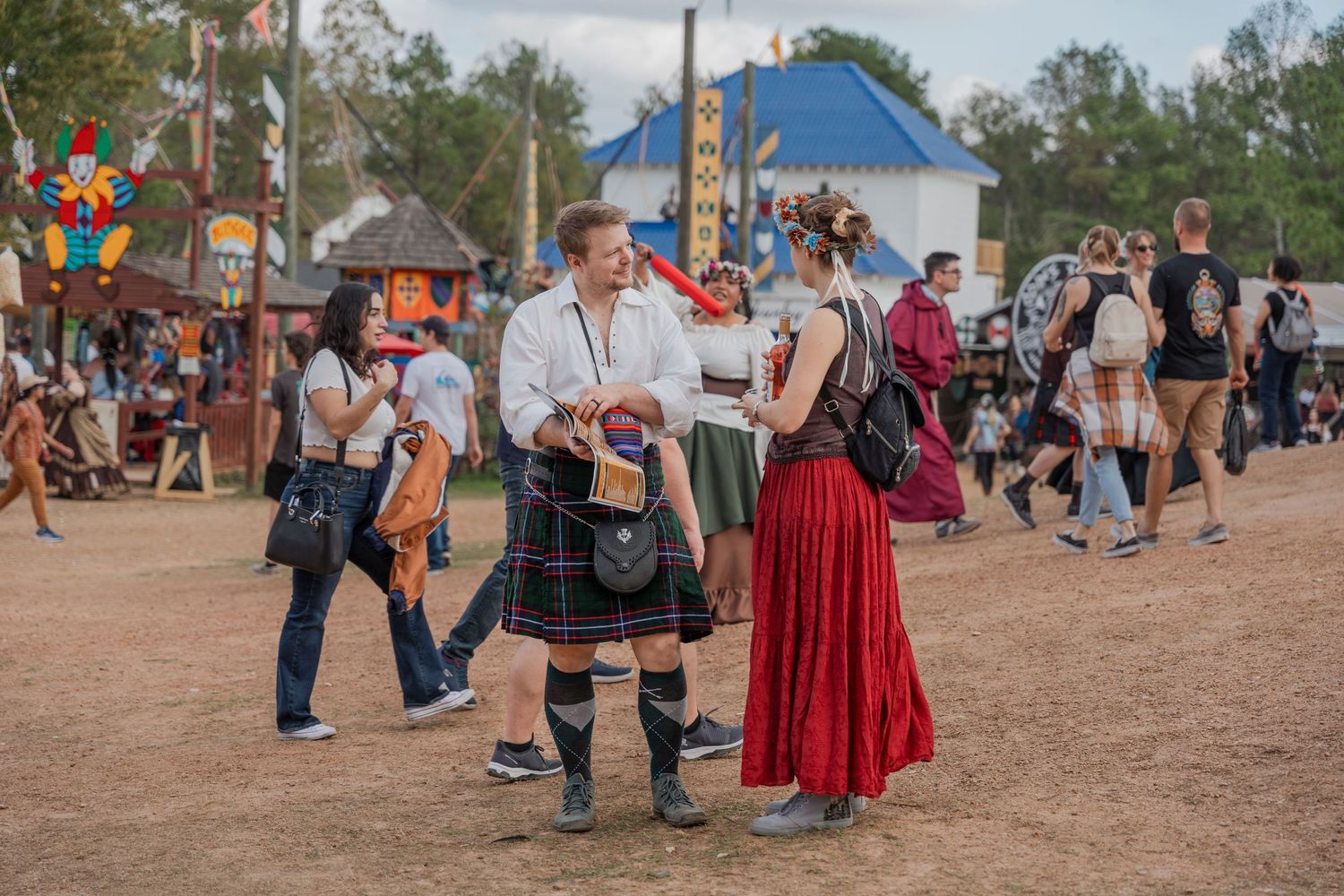 Attendees in traditional Scottish and Renaissance attire gather at a festival on dusty grounds.