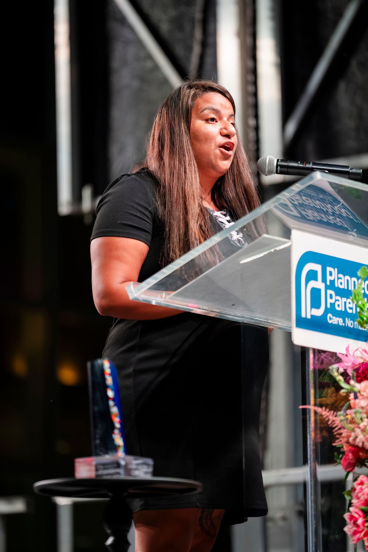 A speaker in a black shirt delivers remarks at a podium during an outdoor event.