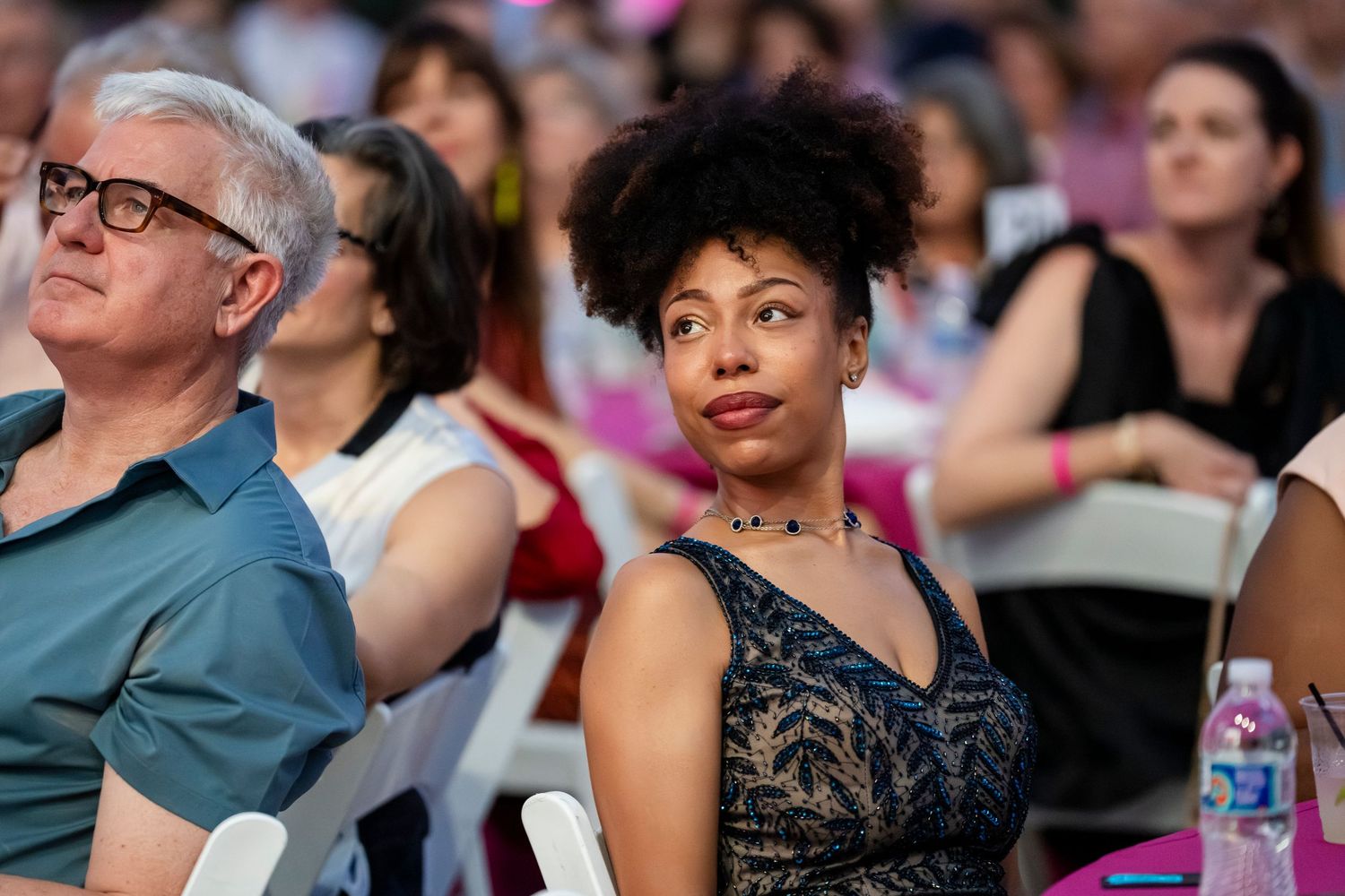 Audience members watch attentively during an event, with people seated in rows wearing casual summer attire.