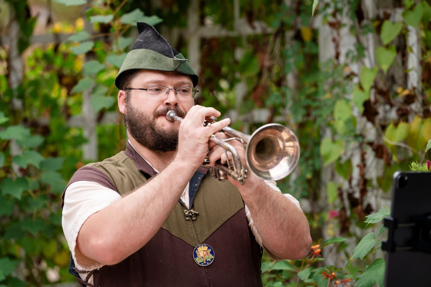 A musician plays a brass trumpet outdoors against a green leafy background.