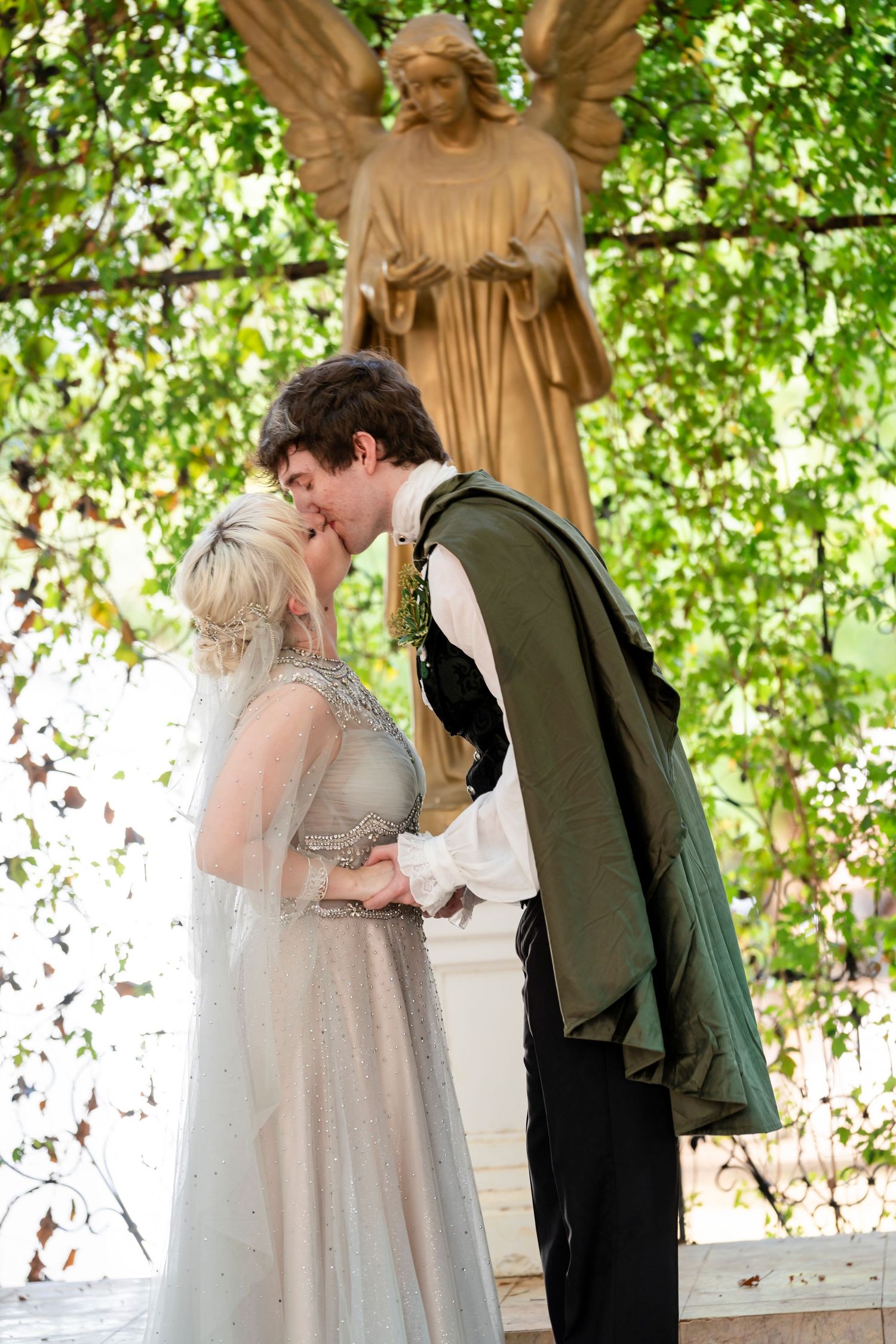 A couple shares a romantic kiss under a canopy of green leaves and stone statues at their wedding ceremony.