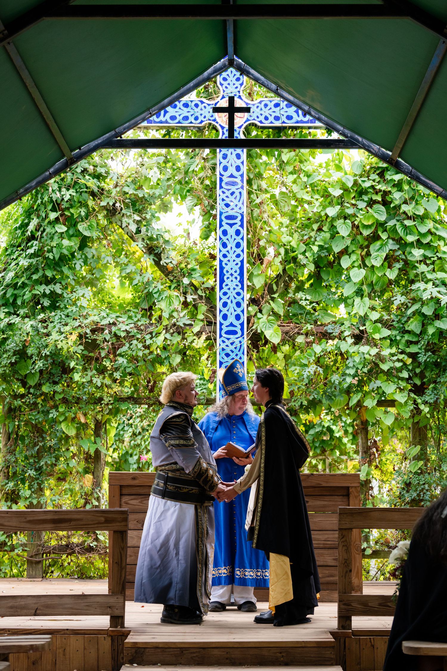 A couple stands at an outdoor wooden gazebo with stained glass and cross details during a wedding ceremony.