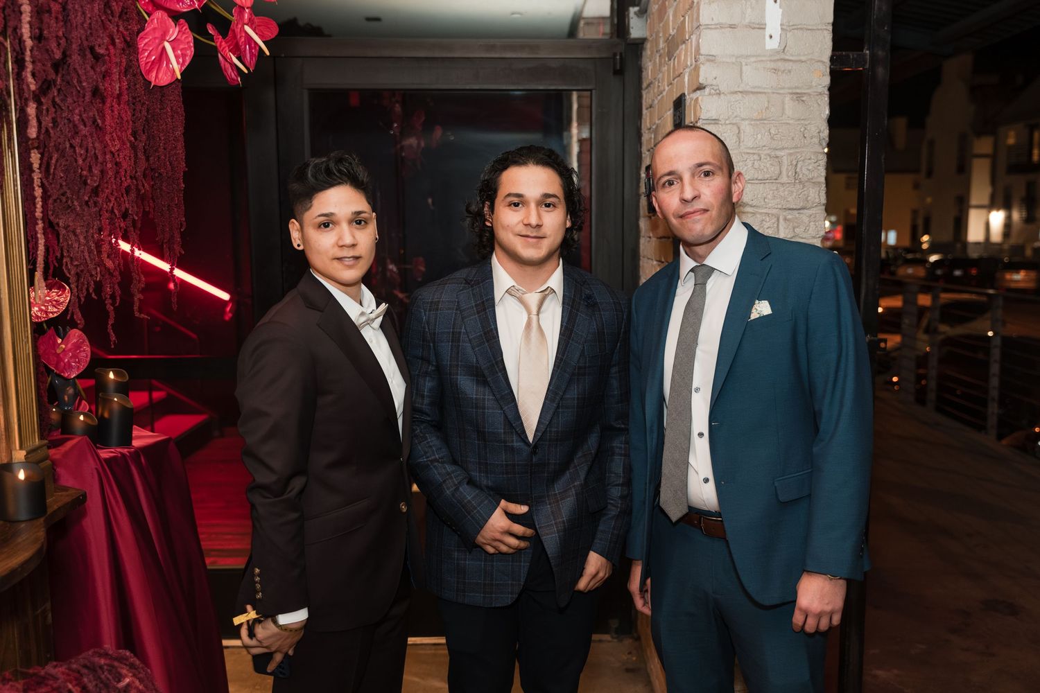 People in formal attire pose together at an evening event with red lighting and brick walls in the background.