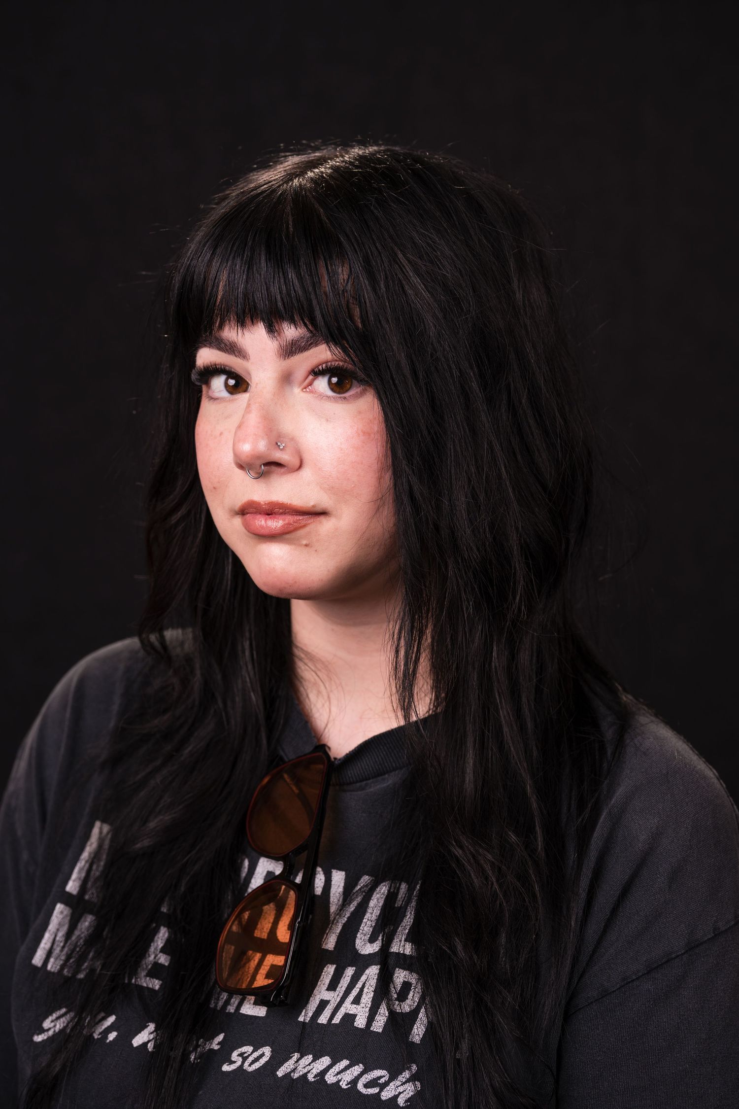 Person with dark hair and dramatic makeup poses against black background wearing a black hoodie with text.