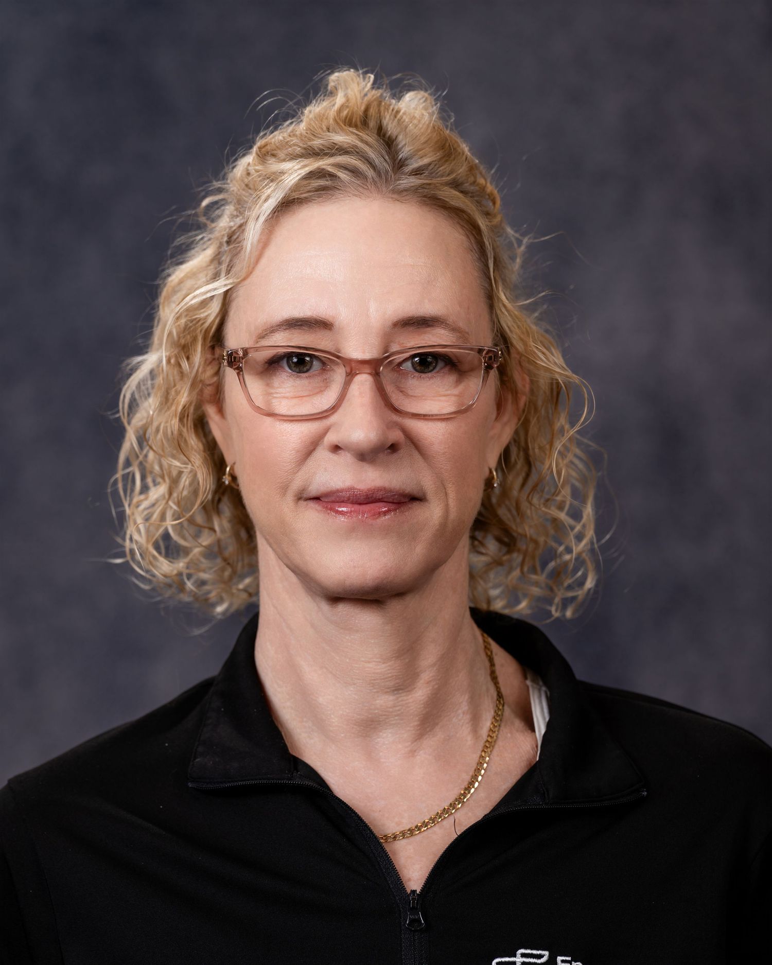 Professional headshot of someone with curly blonde hair and glasses wearing a black top against a dark background.