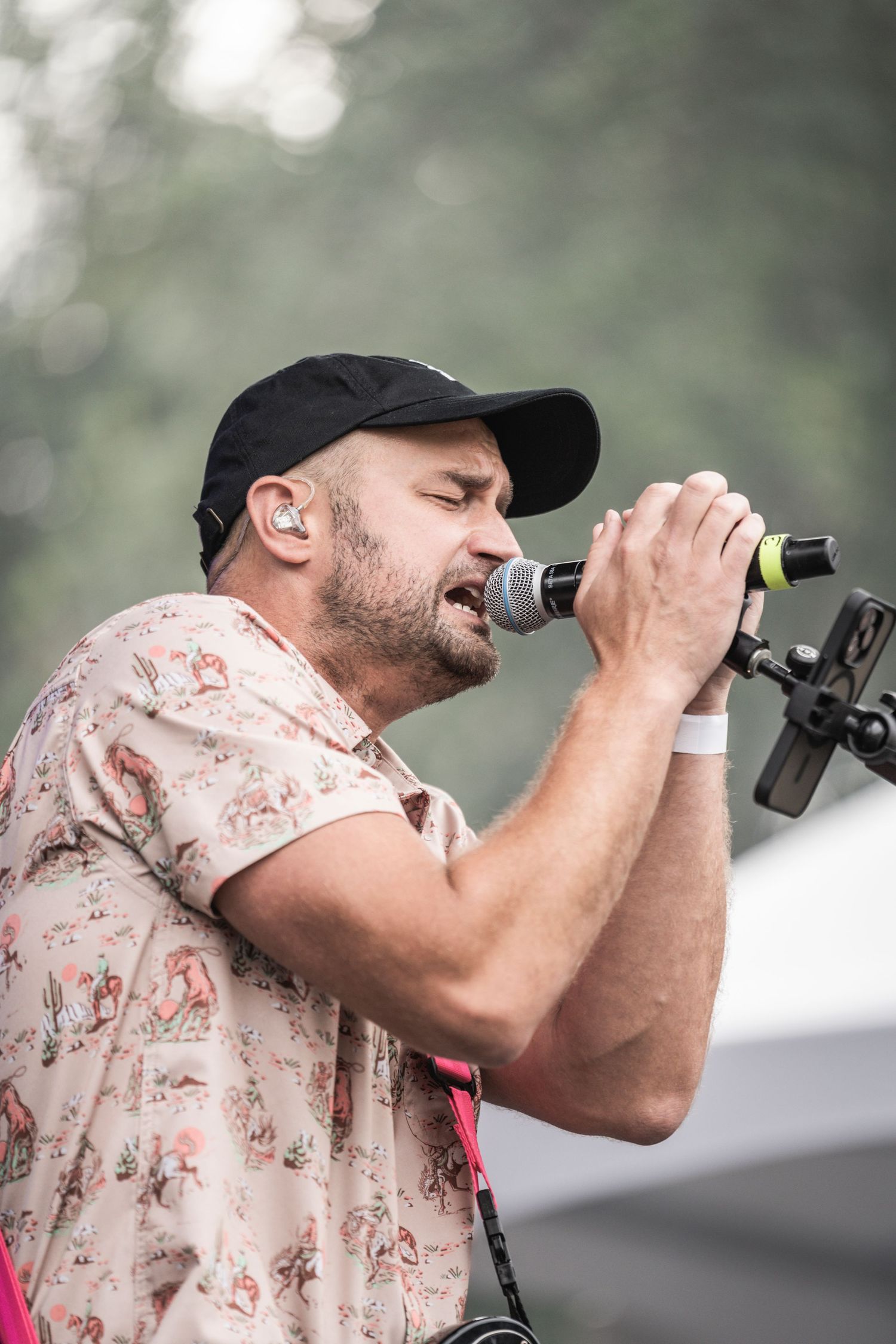 Singer in floral shirt and baseball cap performs passionately on stage with microphone at outdoor concert venue.